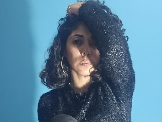 Lola Petterson, con i capelli neri ricci, indossa un maglione nero scintillante, orecchini a cerchio e una collana d'argento, con una mano tra i capelli, sguardo serio, sfondo blu, viso leggermente in ombra..
