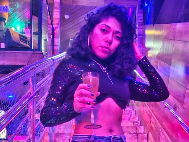 Lola Petterson, con i capelli ricci, indossa un top corto nero scintillante, tiene un cocktail in mano e si trova su una scala illuminata da luci al neon, con un'espressione seria. Visibili il seno, la parte inferiore dell'addome e il cocktail in mano..