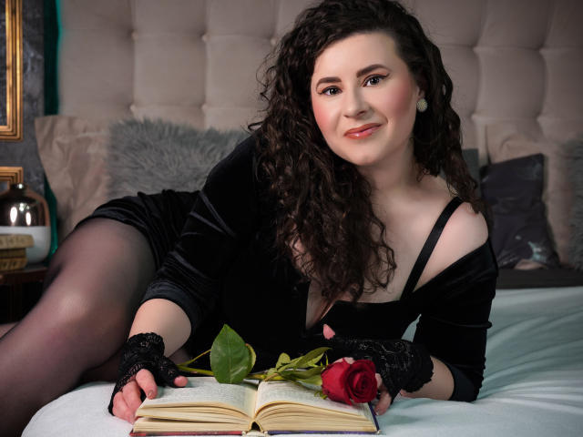 GorJesHot mit lockigem, dunklem Haar, in einem schwarzen Kleid, mit einem mit Rosen bedeckten Buch auf dem Bett, trägt schwarze Spitzenhandschuhe, lächelt in die Kamera, zeigt ihre Brüste und ihren Hintern leicht, liegt auf weißen Laken..