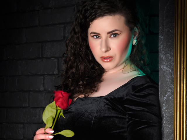 GorJesHot with curly dark hair, holding a red rose, wearing a black velvet dress, standing in a dark brick wall, golden frame on the right.