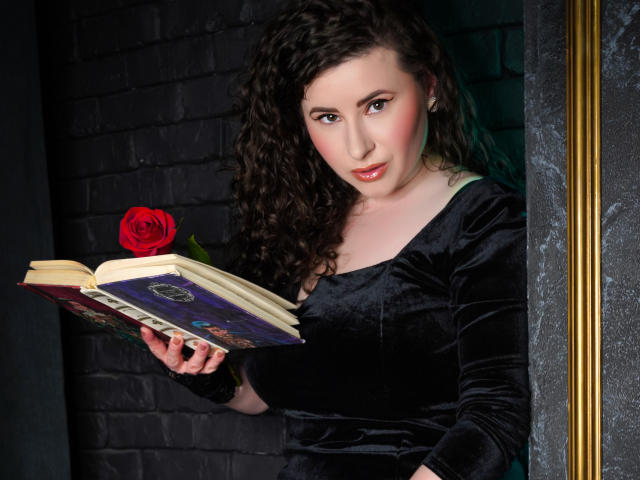 GorJesHot, con i capelli ricci e scuri, indossa un abito di velluto nero, tiene in mano un libro aperto con una rosa rossa, ha un viso attraente, di carnagione chiara e labbra rosate..