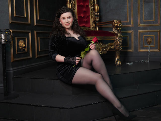 GorJesHot, con cabello castaño rizado, vestido de terciopelo negro, guantes de encaje y medias transparentes, sosteniendo una rosa, sentada en escalones oscuros, con las piernas cruzadas, y una silla roja con detalles dorados detrás..