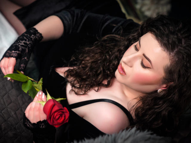 GorJesHot, con cabello castaño rizado, guantes de encaje negros y una rosa roja, sosteniéndola cerca de sus pechos, vestida de negro, con los ojos cerrados, luciendo sexy y seductora..
