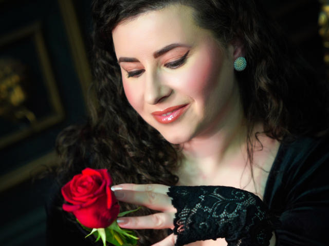 GorJesHot with curly brown hair, holding a red rose, wearing black lace glove, and sparkling earrings, smiling with pink lips, looking down at rose.
