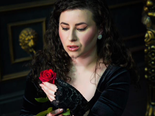 GorJesHot with curly dark hair, holding a red rose, wearing a black velvet dress with lace sleeves, looking down with pink lips, pale skin, and diamond earrings.
