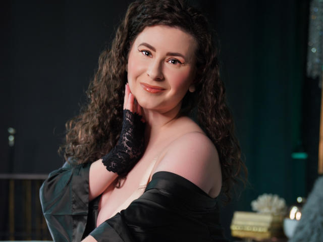 GorJesHot, avec ses cheveux bruns bouclés, souriante, portant un gant en dentelle noire sans doigts et une robe en satin noir à épaules dénudées, laissant apparaître ses épaules et sa poitrine. Éclairage doux et chaleureux..