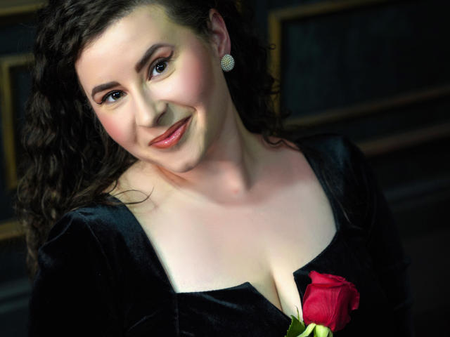 GorJesHot, avec ses cheveux noirs bouclés, ses joues roses et sa robe en velours noir, tenant une rose rouge, souriant à l'appareil photo, montrant ses seins, portant des boucles d'oreilles en diamants, dans une pièce sombre..