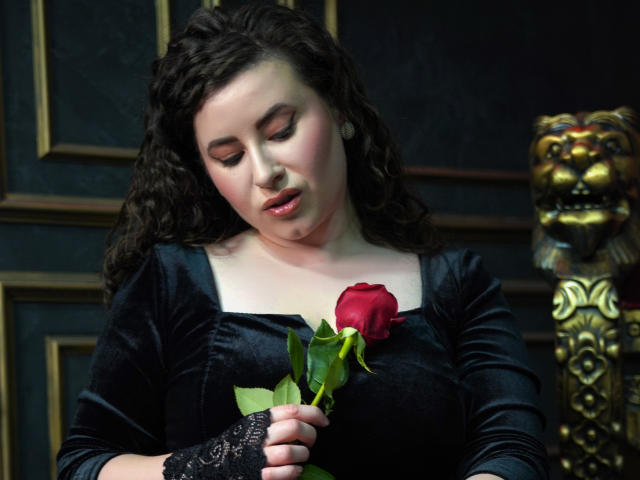 GorJesHot, avec ses cheveux bruns bouclés, portant une robe en velours noir, tenant une rose rouge devant ses seins, gant en dentelle noire sans doigts, regardant vers le bas avec les lèvres légèrement entrouvertes..