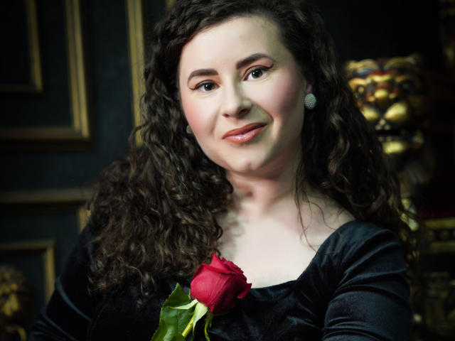 GorJesHot, con cabello castaño rizado, vestida con un vestido negro de terciopelo, sosteniendo una rosa roja, sonriendo y con aretes brillantes. Se ven los senos, pero no son el foco principal..