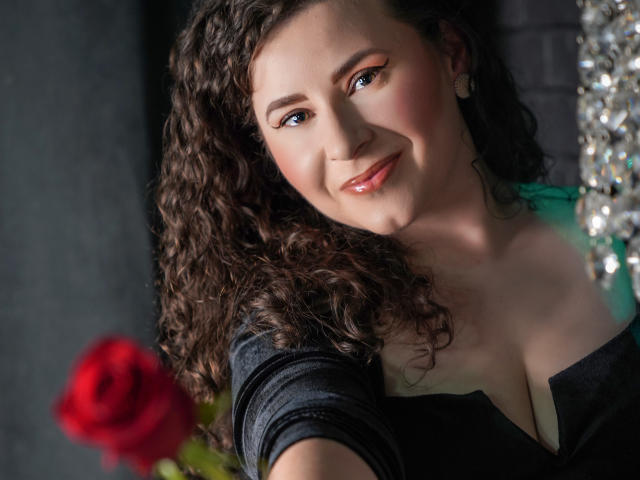GorJesHot, con i capelli castano ricci, gli occhi azzurri e le labbra rosa lucide, sorride a una rosa rossa in primo piano, indossando una maglia nera che mette in risalto il décolleté..