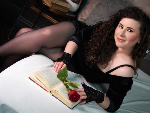 GorJesHot with curly brown hair, black dress, and lace gloves, laying on bed, legs crossed, holding red rose over open book, smiling, wearing sheer black pantyhose.