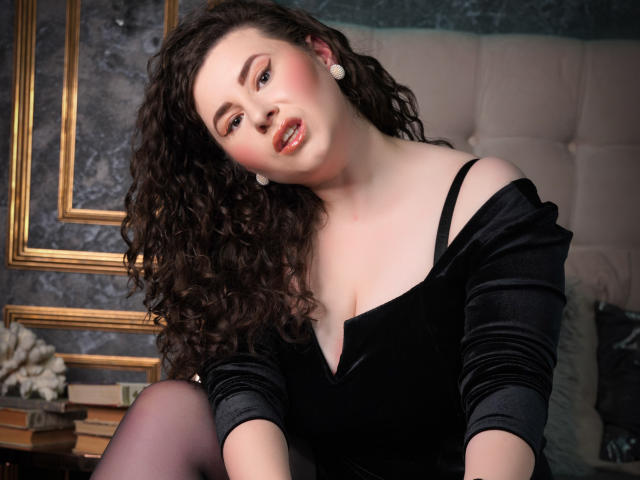 GorJesHot has curly brown hair, fair skin, and wears a black off-shoulder dress with white earrings, showing her boobs and a slight smile in a cozy room.