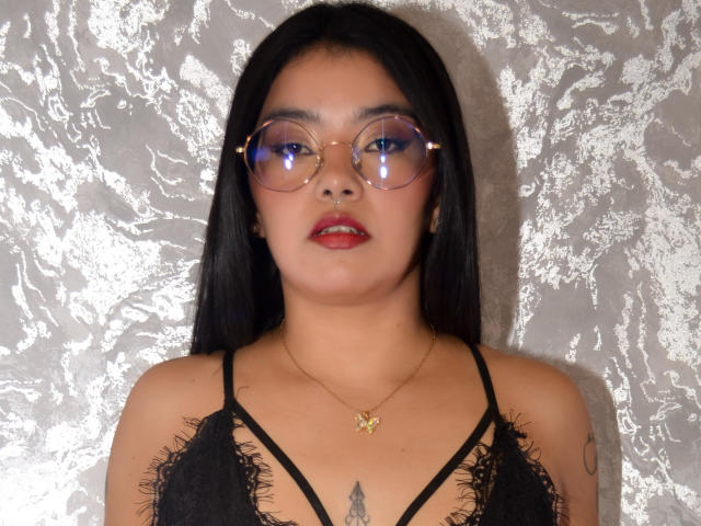 Emma Ruiz, con cabello negro largo, usando gafas redondas, lápiz labial rojo, una blusa de encaje negro, un collar dorado y un tatuaje visible debajo de sus senos, frente a una pared plateada con textura..