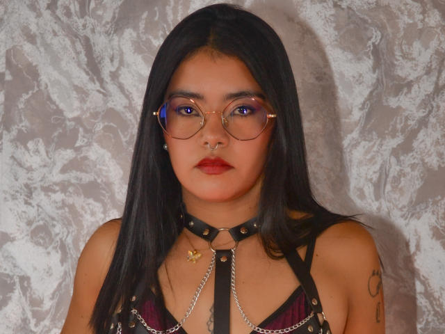 Emma Ruiz, met zwart haar, een bril, een neusring, rode lippenstift, een leren harnas, kettingen, een tatoeage op haar rechterarm, kijkt naar de camera, met een grijze, gestructureerde muur op de achtergrond. Haar borsten zijn zichtbaar in het harnas..