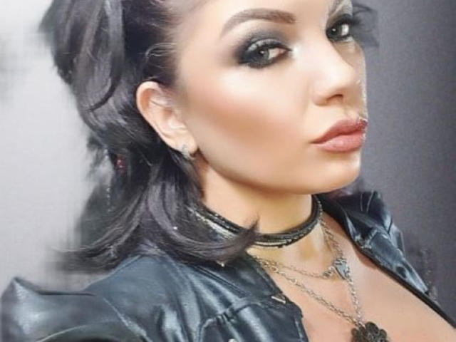 CristinAnn, con el cabello oscuro recogido en una coleta, vestida con cuero negro y collares, tiene labios brillantes, delineador de ojos marcado y un collar de cadena, y se muestra en un selfie mostrando sus senos..