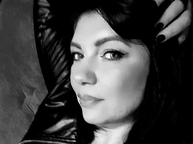 Black-and-white photo of CristinAnn with dark hair, subtle makeup, and black nail polish, lying back with a leather arm, looking slightly to the side with a relaxed expression.
