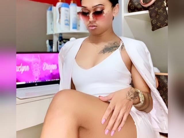 Baddie24 con occhiali da sole rosa, canotta bianca e pantaloncini bianchi, seduta con le gambe incrociate, che mostra il sedere, con tatuaggi sul petto e sul braccio e con le unghie rosa..