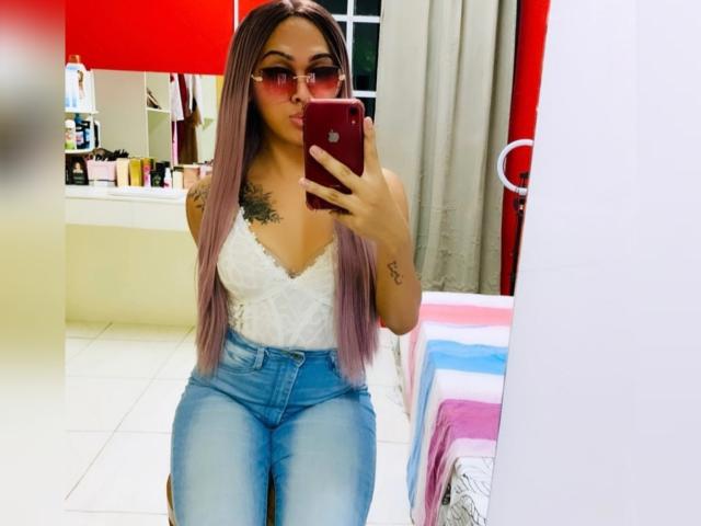 Baddie24, avec de longs cheveux rose-brun, des lunettes roses, un haut en dentelle blanche et un jean bleu, prend un selfie devant un miroir avec un téléphone rouge, montrant des tatouages et une petite poitrine..