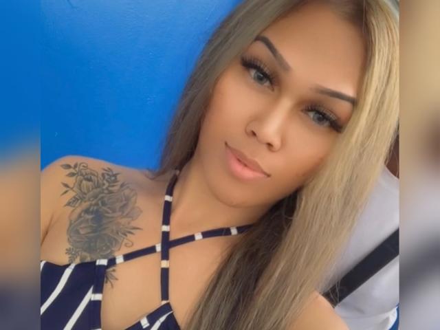 Baddie24, con lunghi capelli biondi, occhi azzurri e un tatuaggio di una rosa sulla spalla destra. Indossa un top a spalline blu a righe e guarda la fotocamera con un leggero sorriso..