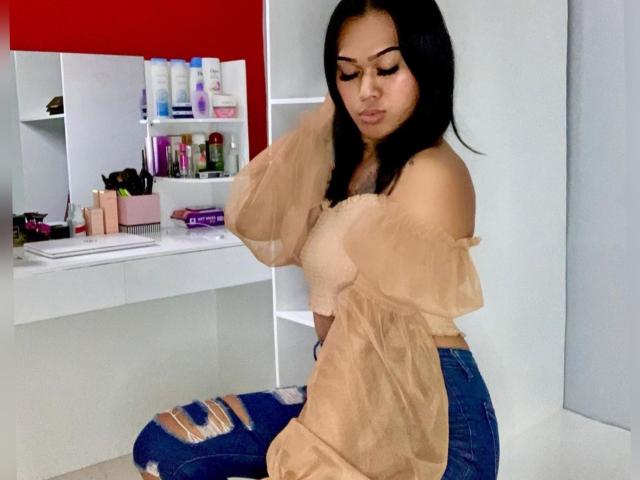 Baddie24 con capelli neri, una maglietta beige a spalle scoperte e jeans blu strappati, seduta, che si tocca i capelli, con lo sguardo rivolto verso il basso, scaffali bianchi con prodotti di bellezza sullo sfondo..