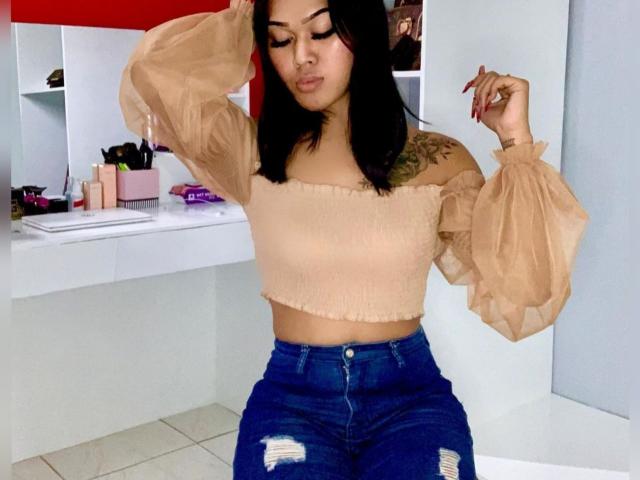 Baddie24 con seni di media grandezza, top color carne a spalle scoperte, maniche a sbuffo, jeans blu, capelli neri, tatuaggio sulla spalla destra, unghie rosse, in piedi in una stanza, con una mano tra i capelli..