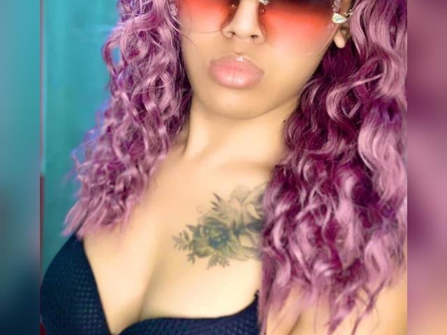 Baddie24, avec ses cheveux bouclés violets, ses lunettes de soleil teintées de rouge, son soutien-gorge noir, et son tatouage de rose sur la poitrine droite, avec des lèvres boudeuses et une peau brun clair..