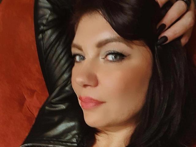 CristinAnn with dark brown hair, black eyeliner, and pink lipstick, wearing a black leather jacket, resting her hand on her head, showing black nail polish.