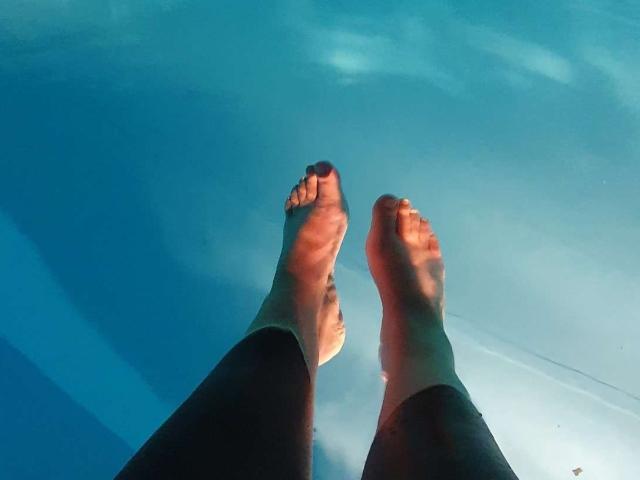 CristinAnn's feet with purple-painted toenails floating in a blue-tinted pool, legs slightly apart, sunlight casting shadows.