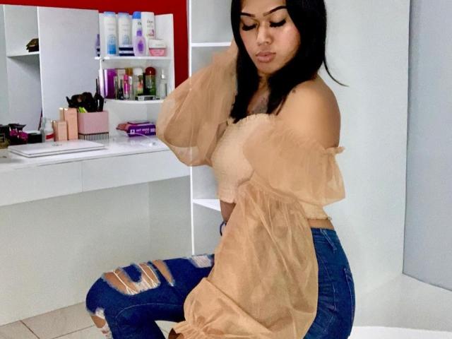 Baddie24, con i capelli neri, indossa una top beige trasparente con una spalla scoperta e jeans blu strappati, è seduta e si sistema i capelli, mostrando il suo seno di medie dimensioni e un viso carino..