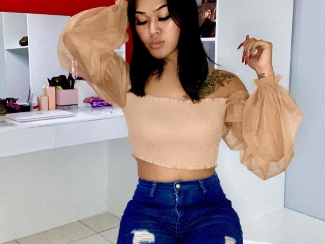 Baddie24 indossa una maglietta beige a spalle scoperte, con maniche a sbuffo, e jeans blu strappati. Ha i capelli neri, un tatuaggio sulla spalla sinistra e sta toccando i capelli con entrambe le mani..