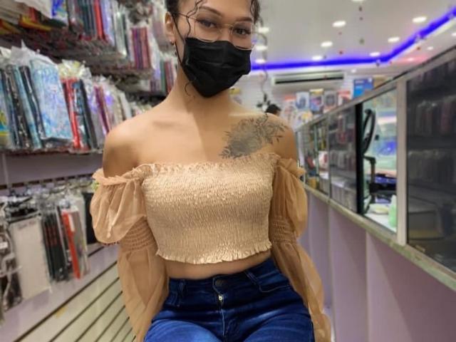Baddie24 with small tits, off-shoulder beige top, blue jeans, black mask, tattoo on right shoulder, standing in a store, wearing glasses, short brown hair, casual look.