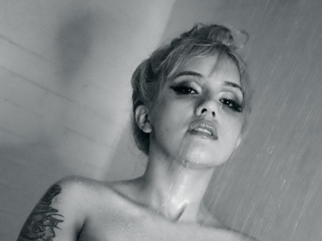 SabrinaCole est nue, allongée sur le comptoir d'une salle de bain, les jambes en l'air, montrant ses seins brillants et son vagin. Elle a des tatouages, les cheveux blonds attachés en chignon, et un sourire sexy..