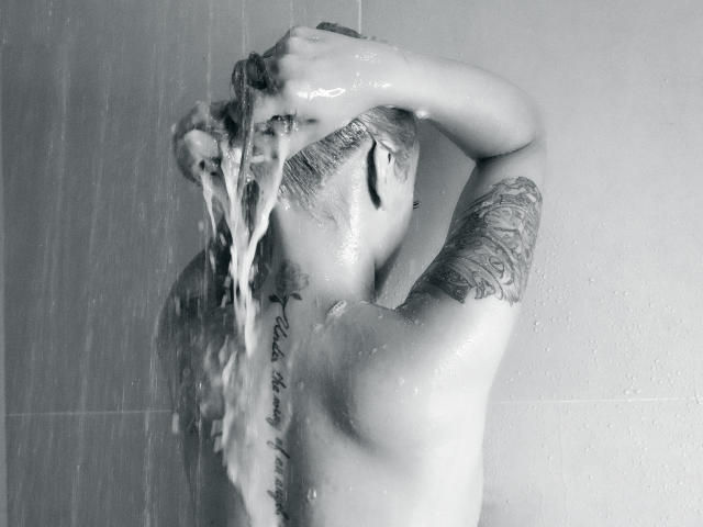 CatalinaJH, avec ses cheveux sombres, portant une veste blanche, de la lingerie en dentelle noire et des bas résille, se tient près d'un tableau blanc avec un dessin de sextoy, sa main posée sur sa hanche..