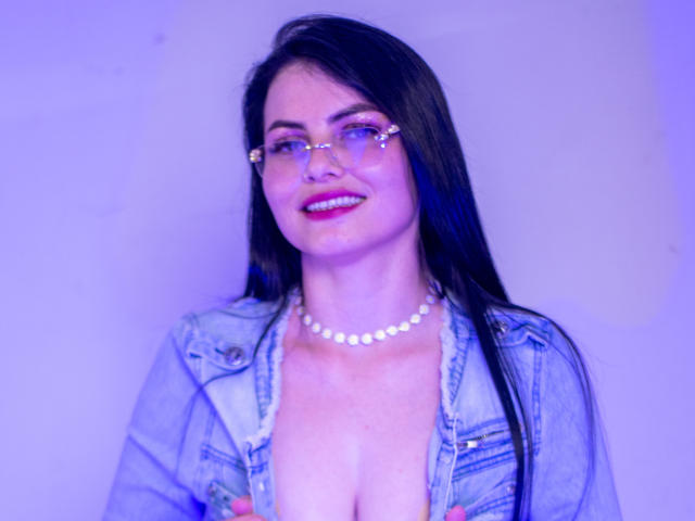 Laurell Charming, con cabello negro, vestida con un vestido blanco de lunares, medias negras y tacones altos negros, chupando una piruleta, con las piernas levantadas, mostrando su vulva con un juguete sexual rosa..
