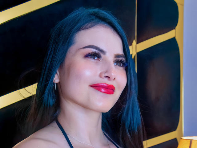 LaurieJemin, avec ses longs cheveux noirs, son rouge à lèvres rouge, son soutien-gorge en dentelle noire et son nœud rouge à pois, regarde vers le haut avec un léger sourire, montrant sa poitrine et un collier blanc épais..