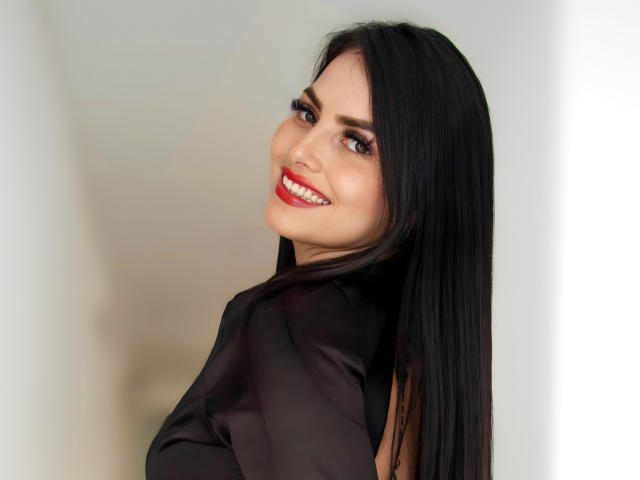 LaurieJemin with black hair, red lipstick, and shiny black top, smiling at the camera, showing white teeth, and looking over her shoulder. She's cute and sexy.