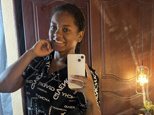 TiffanySammy with dark skin, smiling, holding a white iPhone, wearing a black "GEMV" patterned shirt, touching her chin, with curly hair, small tattoo on right forearm.