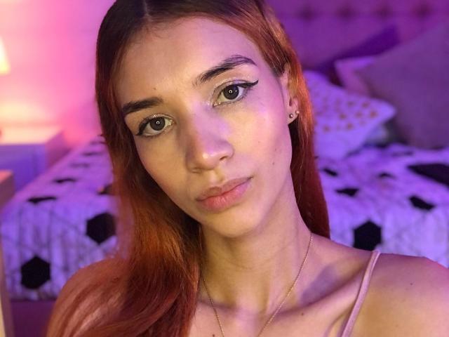 GretaHarris a les cheveux roux, les yeux verts et un joli visage avec un maquillage discret. Elle porte un haut rose et un collier fin. Ses seins sont petits, et elle regarde l'appareil photo..
