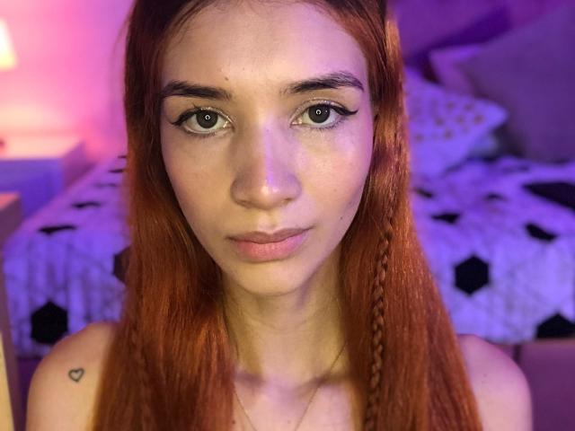 Greta Harris, con cabello rojo, ojos verdes y un pequeño tatuaje de corazón en el hombro derecho. Está mirando directamente a la cámara con una expresión neutra. Fondo: dormitorio..