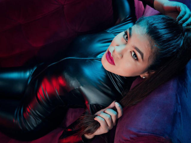 AmyXPlata in einem glänzenden schwarzen Latex-Body, liegt auf einem violetten Sofa, hält ihr langes braunes Haar, trägt roten Lippenstift und hat einen verführerischen Blick, und schaut nach oben in die Kamera..