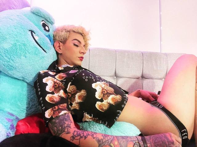 Uma mulher com cabelo descolorido, tatuagens, camisa preta com estampas de donuts e roupa íntima preta, apoiada em um urso de pelúcia azul gigante, deitada em um sofá cinza, com uma expressão relaxada..