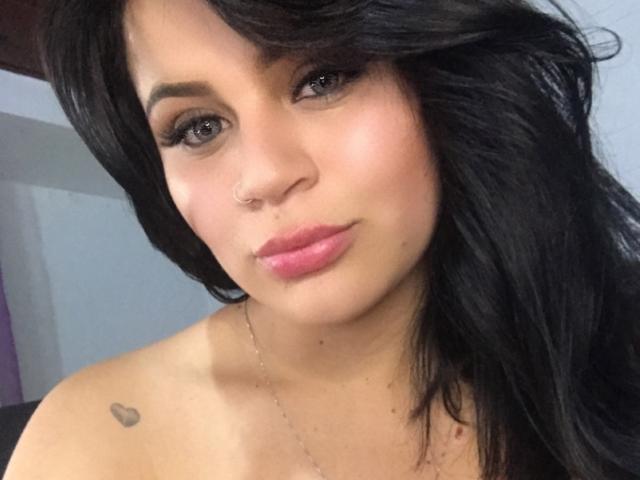 ShantallParis has dark wavy hair, brown eyes, pink lipstick, a nose ring, and a small heart tattoo on her left shoulder. She's wearing a delicate necklace.