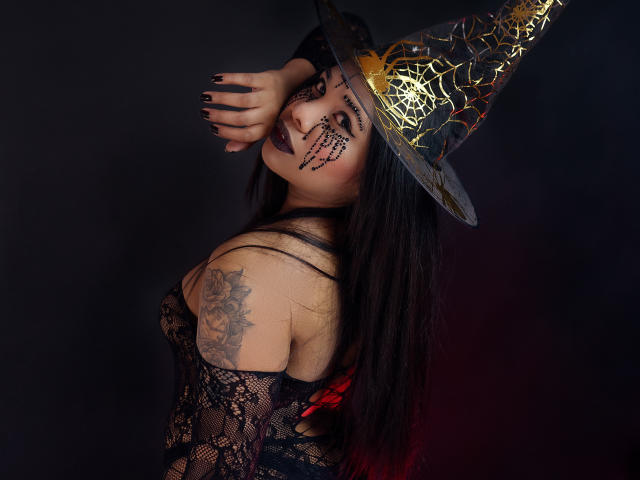 TriniShely met een outfit van zwart kant, gezichtsschildering in de vorm van een spinnenweb, een gouden hoed, lang zwart haar, een tatoeage op de rechter schouder, donkere nagels, profiel, naar boven kijkend, donkere achtergrond..