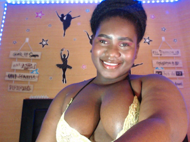 Dilay with dark skin, short curly hair, smiling, wearing a yellow lace bra, showing her big tits, standing in a room with blue LED lights.