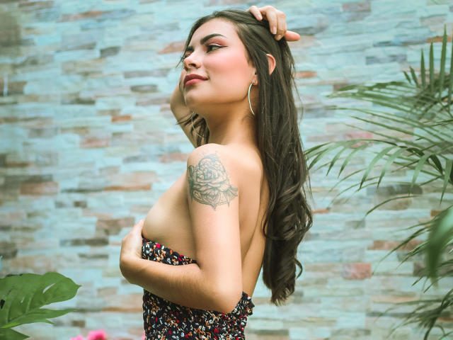 Zenia Murphy, con lunghi capelli scuri, tiene un braccio sopra i suoi seni nudi, mostrando un tatuaggio di una rosa sulla spalla, indossa una gonna floreale e si trova di fronte a un muro di pietra..