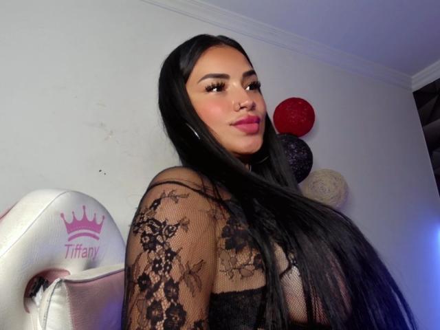 OnniOconor with long black hair, wearing a black lace top, showing her titts, looking at the camera with pink lipstick, a white Tiffany gaming chair on the left.