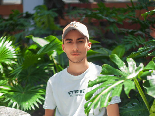 AlexOconor, kurze, braune Haare, helle Haut, trägt eine weiße Mütze und ein weißes Hemd, steht vor grünen Monstera-Blättern und blickt mit neutralem Gesichtsausdruck in die Kamera..