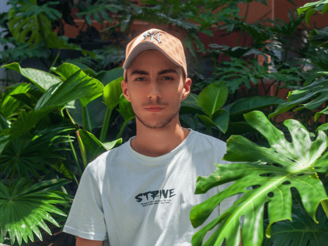 AlexOconor, avec sa peau mate, ses sourcils foncés et sa barbe soignée, porte une casquette beige et un t-shirt blanc, entouré de feuilles vertes luxuriantes, et regarde directement l'appareil photo..