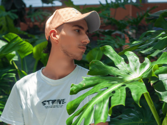 AlexOconor mit brauner Kappe, weißem T-Shirt und Seitenbart, blickt auf ein großes, grünes Monstera-Blatt. AlexOconors Gesicht ist leicht gedreht und zeigt das Profil. Sonnenlicht beleuchtet das Blatt und AlexOconors Haut..