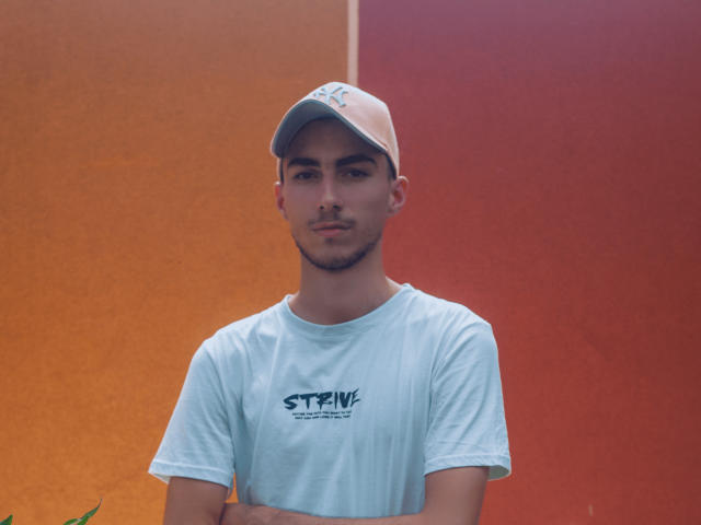 AlexOconor, met een beige pet en een lichtblauwe shirt, staat met armen over elkaar, met een neutrale uitdrukking en een gladgeschoren gezicht, voor een warme oranje en rode muur..